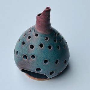 May include: A bulbous ceramic vessel with a teal and reddish-brown glaze, featuring numerous circular perforations across its rounded body and a single protruding spout at the top. The base is flat and circular with a wide opening at the bottom.