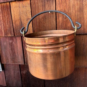 May include: A copper-colored metal bucket with a black handle hangs against a wooden wall. The bucket has a rounded shape with a rolled rim and a sturdy, curved handle. The metal has a warm, reflective sheen.