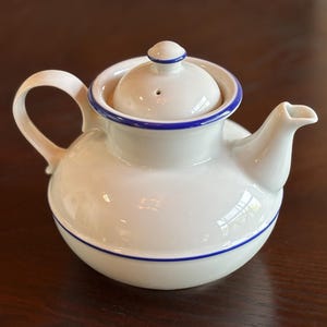 May include: White ceramic teapot with a rounded body, a curved handle, and a spout. The lid and base are accented with a thin blue stripe. The lid has a small knob and a vent hole. A classic design for serving tea.