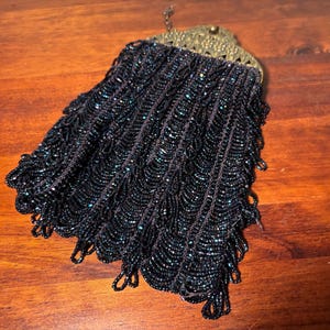 May include: A small black beaded pouch featuring a scalloped pattern of dark iridescent beads. The bag is topped with an ornate, antique-style brass metal frame with a filigree design and a small clasp at the top center for opening and closing.