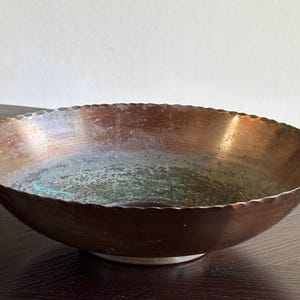 May include: A shallow, round copper bowl with a scalloped edge. The exterior features a warm, metallic copper finish, while the interior displays a weathered, textured surface with visible green oxidation and patina across the bottom basin.