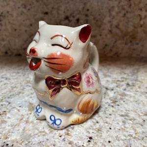 May include: A vintage ceramic cat creamer with a smiling face, red bow tie, and floral accents. The white creamer has red ears, pink cheeks, and a red mouth. Gold trim and blue accents complete the design. A charming collectible.
