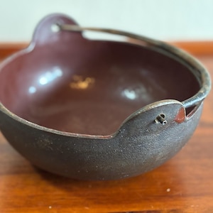 May include: A dark brown cast iron bowl with a burgundy interior. The bowl has a curved handle and two small, rounded protrusions on either side. The bowl is sitting on a wooden surface.