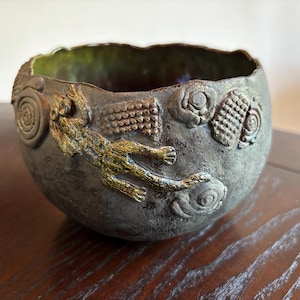 May include: A dark gray ceramic bowl with a textured, uneven rim and a matte finish. The exterior features raised, gold-toned relief decorations including a feline figure, spiral patterns, and rectangular grid motifs. The interior has a green glaze.
