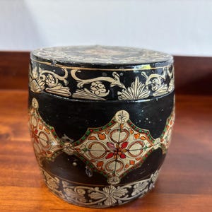 May include: A black cylindrical box with intricate hand-painted designs in gold, red, white, and green. The box has a floral pattern and geometric shapes.