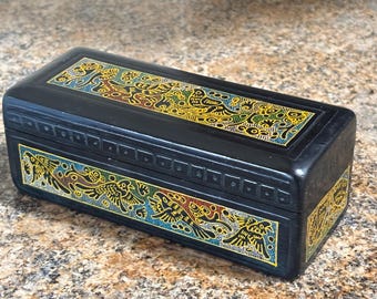May include: A rectangular black wooden box with intricate, colorful designs. The box features detailed patterns in yellow, blue, red, and green, with a repeating bird-like motif. The box is likely used for storage or display.