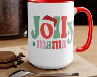 May include: White ceramic mug with a red interior and handle. The mug features the words "Jolly mama" in green, red, and pink lettering, with a Santa hat on the letter "O" and a Christmas ornament on the letter "y".