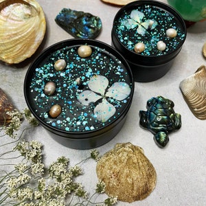 May include: Two black candle jars with teal glitter, white flowers, and pearl accents. The candles are surrounded by seashells and a green turtle figurine.