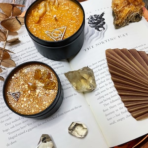 May include: Two golden yellow candles with glitter and small crystals in black metal tins. The candles are on a book with the text "The Sorting Hat" and a brown fan.