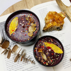 May include: Two black candle jars with a burgundy wax base. The jars are filled with gold glitter, small orange and red crystals, and a dried yellow leaf. The jars are sitting on a book with text about Greek mythology.