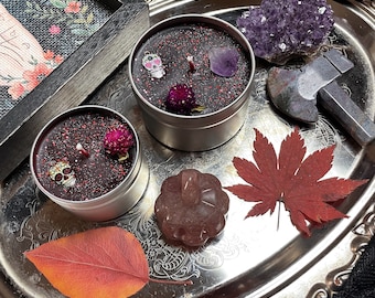 May include: Two round silver metal tins containing black wax candles topped with red glitter, small skull charms, purple amethyst crystals, and dried pink globe amaranth flowers, arranged on a silver tray with a red maple leaf and a pumpkin carving.