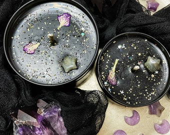 以下が含まれることがあります： キラキラと光る黒いワックスと紫の花のアクセントが付いた黒いキャンドルジャーが2つ。ジャーはアメジストの結晶と紫の星と三日月の形に囲まれています。
