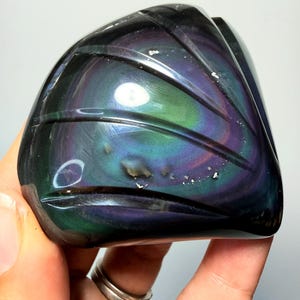 May include: A polished rainbow obsidian stone featuring a smooth, dark surface with iridescent bands of green and purple. The stone is carved with several deep, curved grooves that follow the natural circular patterns of the mineral.