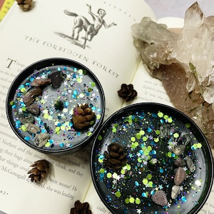 May include: Two black candle tins with dark wax, decorated with stones, glitter, and pine cones, sit on an open book. The book's pages display text and an illustration of a centaur. The candles have a celestial theme.