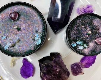 May include: Two teal green and black candles with glitter and dried flowers on a white plate with a purple amethyst crystal and a green crystal.