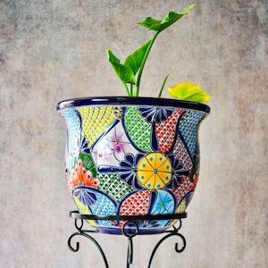 Puede incluir: Un macetero de cerámica pintado a mano, colorido, con un soporte de metal negro. El macetero presenta un patrón geométrico con flores y puntos en tonos de azul, verde, amarillo, rojo y blanco.