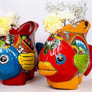 May include: Two colorful ceramic fish planters with white flowers. The fish are painted in bright colors with intricate designs. The planters are perfect for adding a touch of whimsy to any home.
