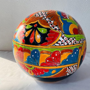 May include: A spherical ceramic object featuring a vibrant, hand-painted design with bold sections of red, blue, yellow, green, and black. The surface is decorated with intricate floral patterns, scalloped borders, and a grid-like lattice motif.