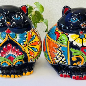 May include: Two ceramic cat figurines featuring glossy black heads with blue eyes and red inner ears. The bodies are decorated with vibrant, multi-colored floral and geometric patterns in yellow, orange, blue, green, and red on a black base.
