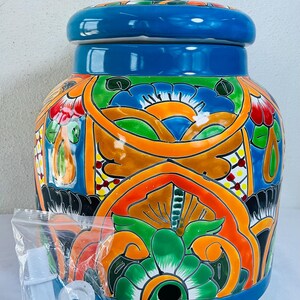 May include: A vibrant, hand-painted ceramic water dispenser with a lid. The jar features a blue base and lid, with intricate floral and geometric patterns in orange, green, and red. A plastic bag of dispenser parts is included.