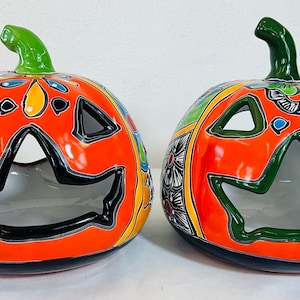 May include: Two ceramic jack-o'-lanterns featuring bright orange surfaces with hand-painted floral and geometric patterns in blue, yellow, and black. Each pumpkin has a green stem and cut-out eyes and mouths with dark outlines.