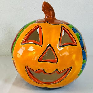 May include: A glossy ceramic jack-o'-lantern featuring a bright orange pumpkin body with a brown stem. The face has triangular eyes and a wide, jagged mouth cutout. The sides are decorated with colourful blue, green, and red floral patterns.