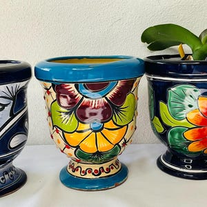 May include: Three colorful ceramic flower pots with floral designs. The pots are blue, white, and yellow with red, green, and orange accents. The pots are all different sizes and shapes.