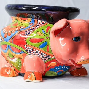 Puede incluir: Una maceta de cerámica con forma de cerdo, con cuerpo rosa y ojos azules. El cuerpo de la maceta está decorado con coloridos diseños florales en rojo, verde, naranja y azul. El borde es azul oscuro.