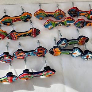 May include: A selection of hand-painted ceramic cabinet pulls in various colours. Each pull has a unique design with vibrant shades of orange, blue, red, and green. The pulls are mounted on metal rods against a white backdrop.