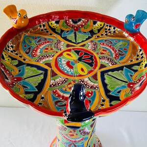 May include: A ceramic birdbath featuring a wide, scalloped bowl with a vibrant, multi-coloured floral pattern in red, yellow, blue, green, and black. Three small, sculpted birds in yellow, blue, and black are perched on the rim of the bowl.