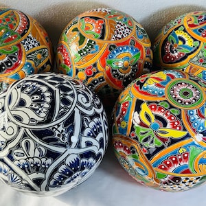 Op de afbeelding: Vijf bolvormige decoratieve keramische objecten met ingewikkelde handgeschilderde patronen. Vier bollen tonen levendige veelkleurige bloem- en geometrische ontwerpen in geel, rood, groen en blauw, terwijl één bol een opvallend marineblauw en wit patroon heeft.