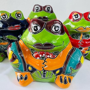 May include: Four ceramic frog figurines arranged in a group. Each frog features a glossy, hand-painted finish with distinct colour combinations including dark blue, bright green, orange, and red, all accented with black outlines and painted lips.