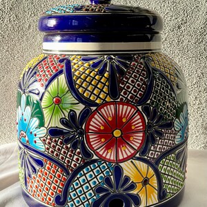 May include: A blue and white ceramic water dispenser with a colorful floral design. The dispenser has a lid and a spout for pouring water.
