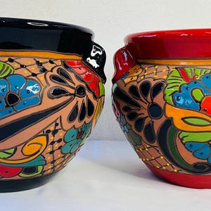 May include: Two ceramic pots with a vibrant floral design. One pot is black, the other is red. Both have handles and are adorned with colorful flowers in red, blue, and green, set against a tan background. The pots are suitable for indoor or outdoor use.