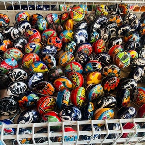 May include: A large collection of round ceramic cabinet knobs featuring a variety of hand-painted floral and geometric patterns in vibrant colors including red, blue, green, yellow, and black, all stored together in a white wire basket.