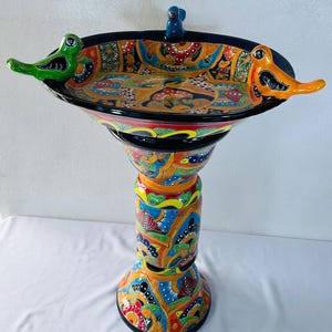 May include: A hand-painted ceramic birdbath featuring a vibrant, multi-coloured floral and geometric pattern. The basin is adorned with three sculpted birds in green, blue, and orange, perched on the black rim, supported by a matching pedestal base.