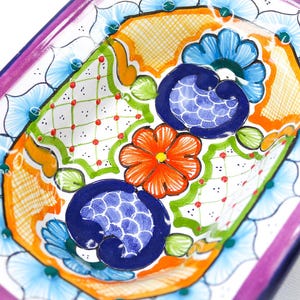 Puede incluir: Un plato de cerámica octogonal colorido con un diseño floral. El plato presenta flores azules, naranjas, verdes y rojas sobre un fondo blanco. El borde del plato está decorado con un borde rosa y azul.