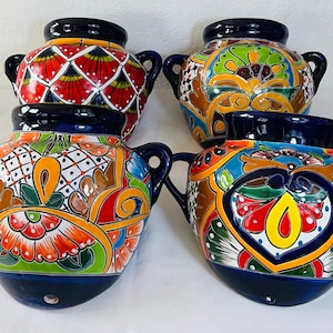 Puede incluir: Cuatro maceteros de pared de cerámica con bordes y asas de color azul oscuro. Cada pieza presenta un diseño único pintado a mano con colores vibrantes que incluyen rojo, naranja, amarillo, verde y blanco, con intrincados patrones florales.