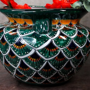 Puede incluir: Un macetero de cerámica verde con un diseño de pluma de pavo real. El macetero tiene una boca ancha y dos asas. El macetero está decorado con pintura verde, blanca, naranja y negra.