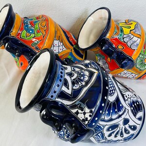 May include: Three ceramic pitchers with wide mouths and side handles. Two pitchers feature vibrant orange, yellow, green, and blue floral patterns. The third pitcher displays a contrasting deep blue and white intricate geometric and floral design.