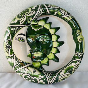 Puede incluir: Una placa de pared de cerámica circular con un diseño de doble cara de sol y luna. La luna es de color crema con detalles pintados en verde, mientras que el sol es de color verde oscuro con rayos de color verde claro. Decorado con motivos florales.