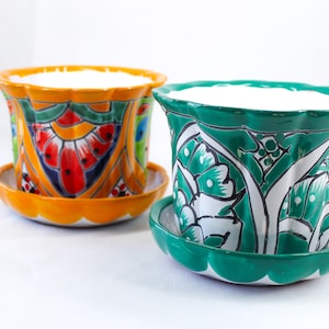 Puede incluir: Dos macetas de cerámica con diseños florales coloridos. La maceta de la izquierda es naranja con flores rojas, amarillas y azules. La maceta de la derecha es verde con flores blancas.