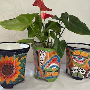 May include: Three colorful, hand-painted ceramic flower pots with geometric designs. The pots are decorated with sunflowers, flowers, and swirls. The pots are sitting on a white surface.