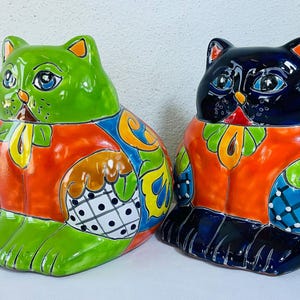 May include: Two ceramic cat figurines. One is green with orange, blue, and yellow accents. The other is black with similar colorful patterns. Both have large eyes and are in a seated position.