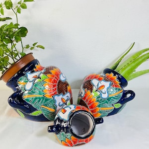 May include: Three glossy, dark blue ceramic pots with two side handles, decorated with vibrant hand-painted orange sunflowers and green leaves. Two pots contain green plants, while the smallest pot is empty and positioned in the front center.