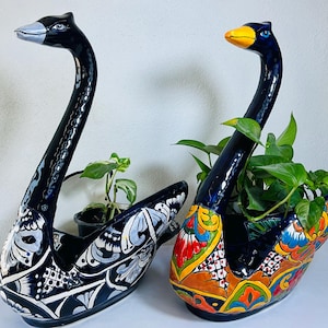 Könnte beinhalten: Zwei Pflanzgefäße in Form von Schwänen aus Keramik. Eines ist schwarz mit weißen und grauen Mustern, das andere ist schwarz mit einem bunten Blumenmuster. Beide haben lange Hälse und sind mit grünen Pflanzen gefüllt. Die Schwäne haben blaue Augen und stehen auf einer weißen Oberfläche.