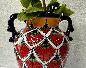 May include: A hand-painted ceramic vase with two handles, featuring a red, white, blue, yellow and green geometric pattern. The vase is filled with a green plant and red flowers.