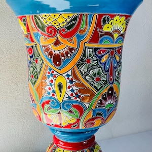 Puede incluir: Una vibrante jardinera de cerámica pintada a mano con base de pedestal. La jardinera presenta un diseño audaz y colorido con intrincados patrones florales y geométricos en rojo, azul, amarillo y verde. El borde y la base son de color azul brillante y contiene plantas verdes.