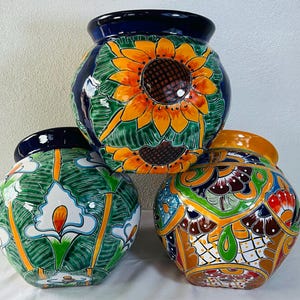 Puede incluir: Tres macetas de cerámica redondas con diseños pintados a mano. La maceta superior presenta girasoles amarillos sobre un fondo verde. La maceta inferior izquierda muestra calas blancas sobre verde, y la inferior derecha tiene un patrón geométrico.