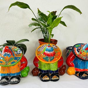 Puede incluir: Tres macetas de cerámica coloridas con forma de personas que llevan sombreros. Las macetas están decoradas con motivos florales y tienen un estilo de arte popular y caprichoso.